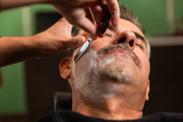 barber shaves a client's cheek full of shaving foam with a razor in a barber shop. the client is tuned into the chair. both caucasian. green, black and brown.